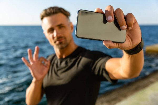 handsome adult man taking selfie with smartphone and showing okay gesture on seashore