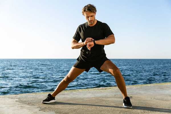 handsome adult sportsman looking at fitness tracker while stretching before training on seashore