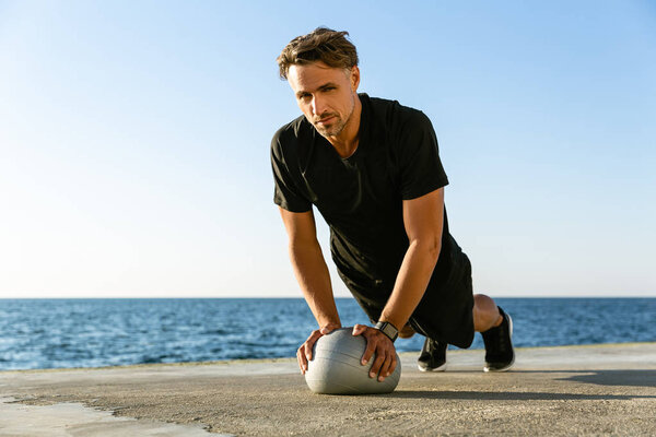 handsome adult sportsman doing push ups with fit ball on seashore and looking at camera