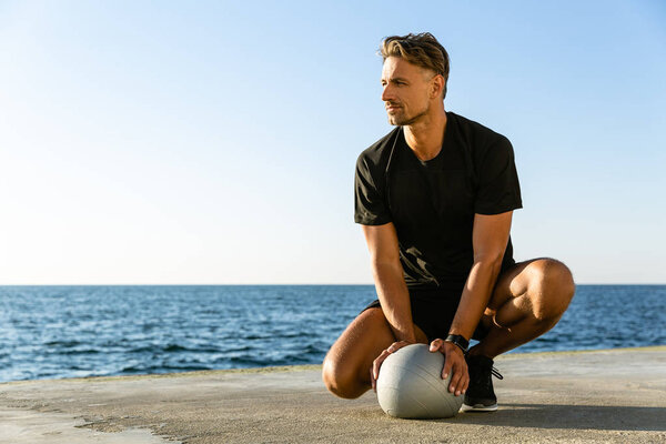 handsome adult sportsman sitting with fit ball on seashore and looking away