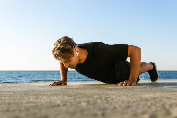 sporty adult man with wireless earphones doing push ups on seashore