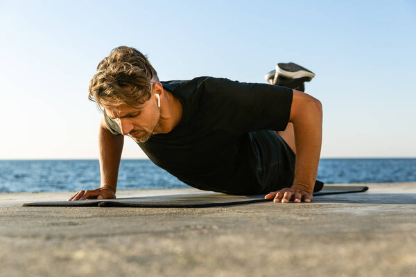 fit adult man with wireless earphones doing push ups on knees on seashore