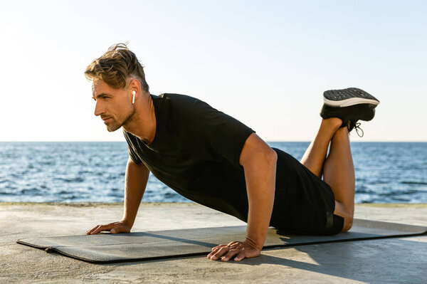 sporty adult man with wireless earphones doing push ups on knees on seashore