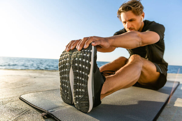 handsome adult sportsman with wireless earphones stretching on seashore