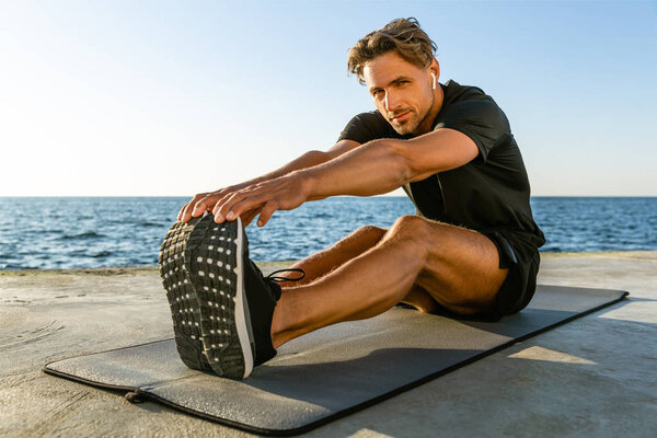 fit adult man with wireless earphones stretching on seashore