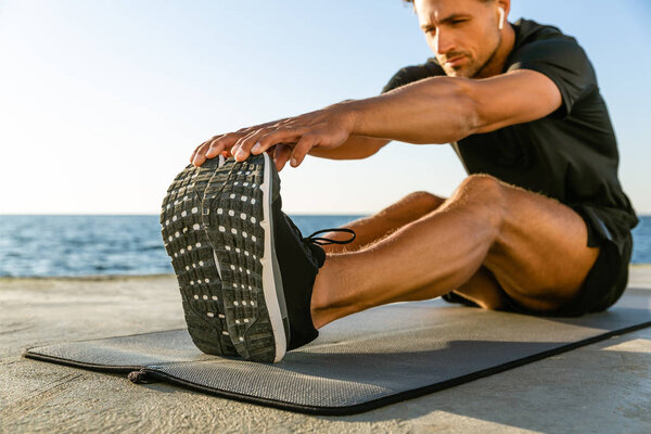 sporty adult man with wireless earphones stretching on seashore