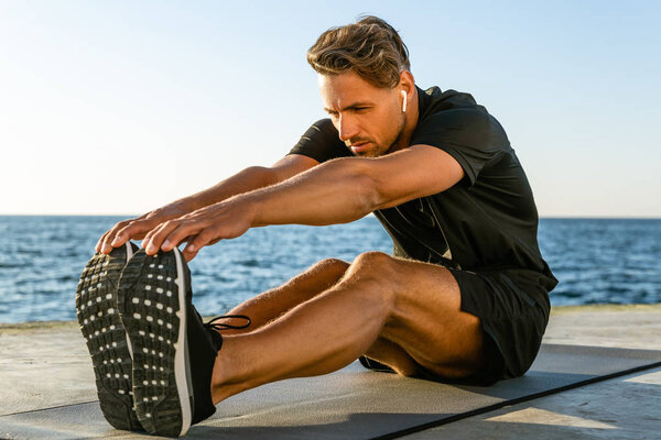 athletic adult man with wireless earphones stretching on seashore
