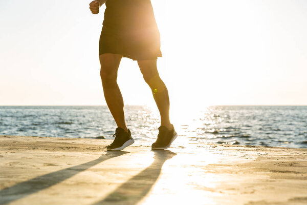 cropped shot of fit man jogging on seashore in front of sunrise