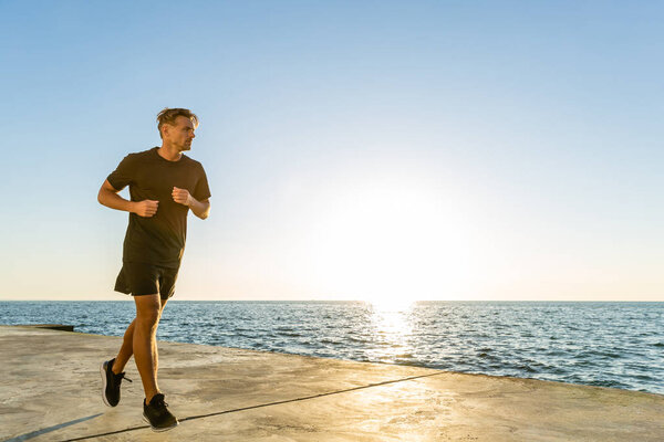 handsome adult sportsman jogging on seashore in front of sunrise