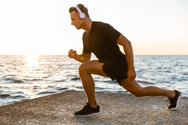 side view of handsome adult sportsman in headphones doing lunges during training on seashore