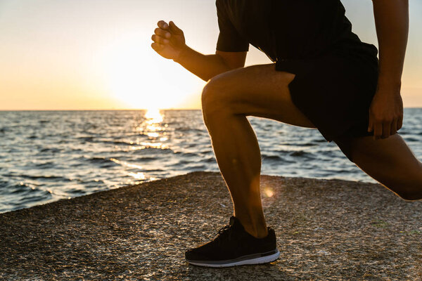 cropped shot of sportsman doing lunges during training on seashore in morning
