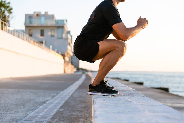 cropped shot of sportsman standing squats on parapet on seashore