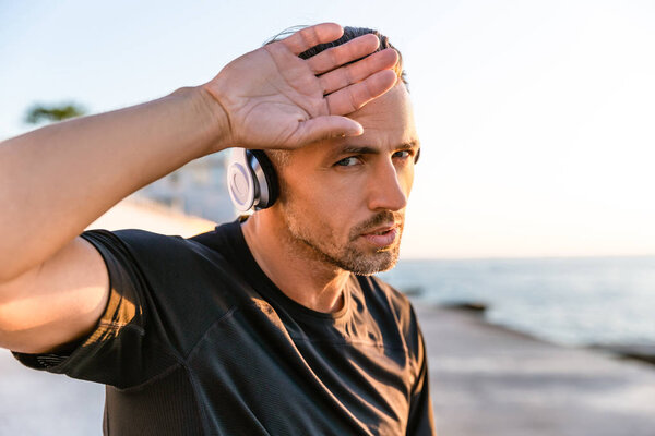 уставший взрослый мужчина в беспроводных наушниках вытирает лоб рукой о берег моря
