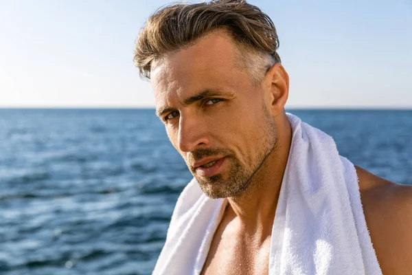 海岸にカメラ目線の肩の上のタオルで上半身裸のハンサムな成人男 ストック画像