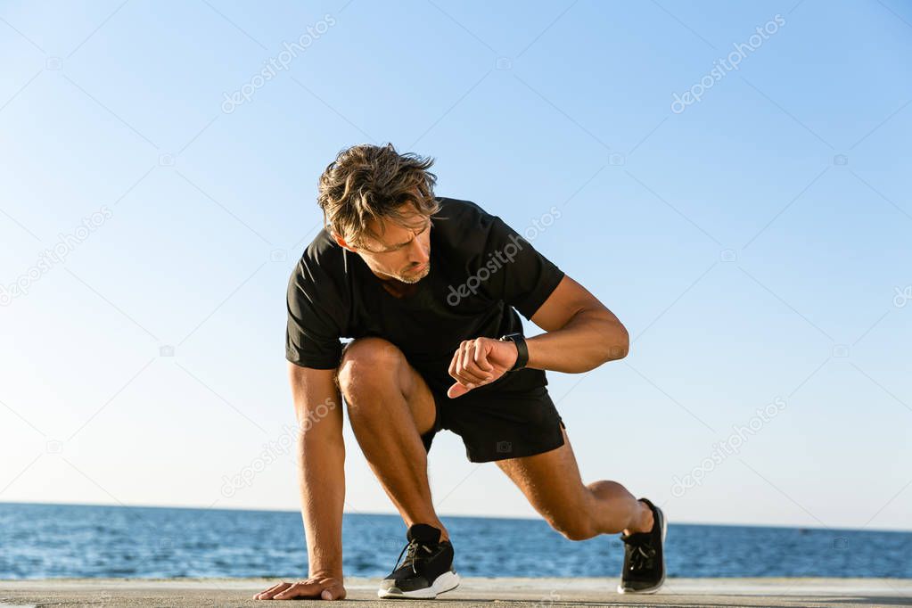 handsome adult sprint runner looking at fitness tracker while standing in start position for run on seashore