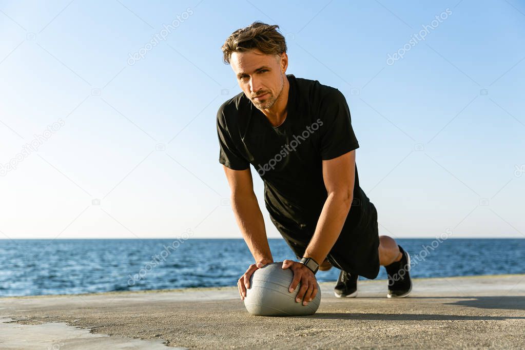 handsome adult sportsman doing push ups with fit ball on seashore and looking at camera