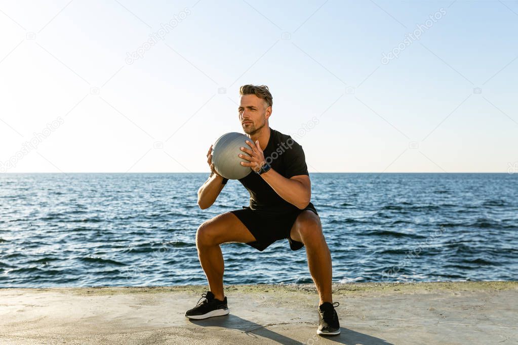 sporty adult man doing squats with fit ball on seashore