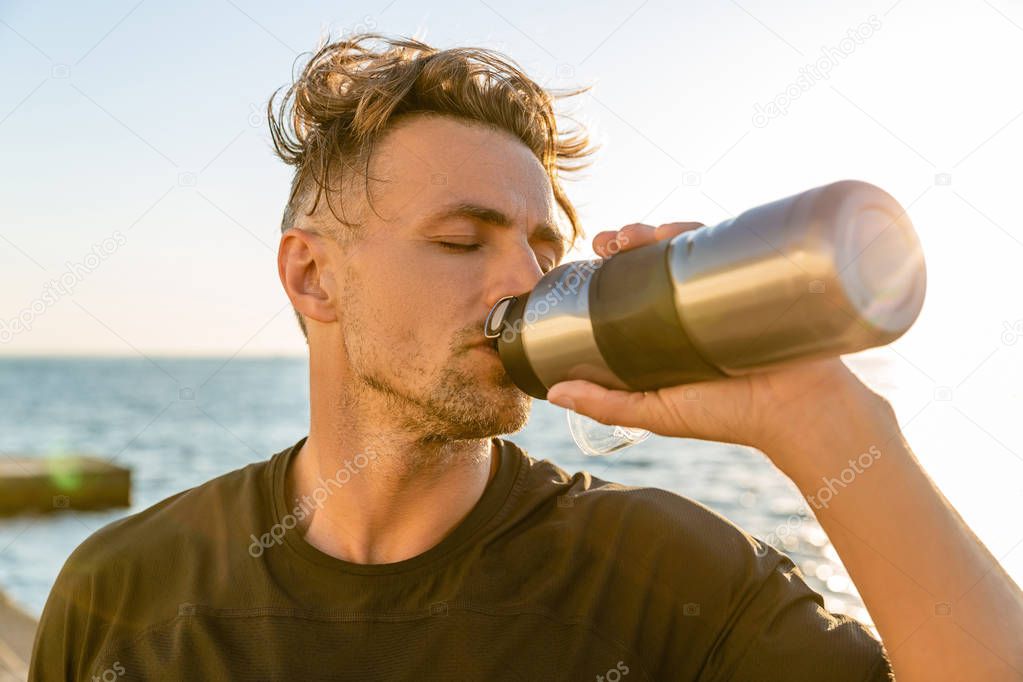 adult sportsman drinking water from fitness bottle on seashore in front of sunrise after workout