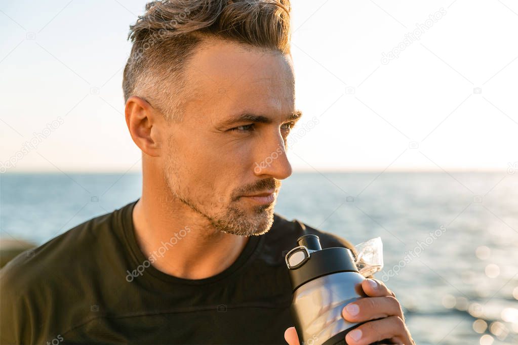 close-up portrait of handsome adult man drinking water from fitness bottle on seashore in front of sunrise