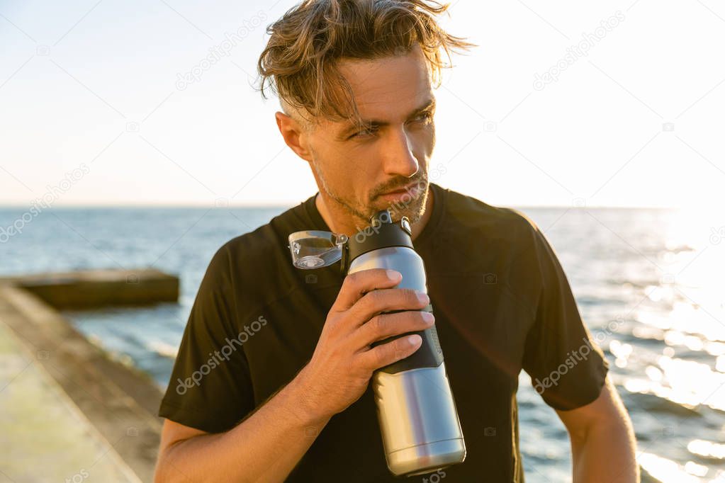 handsome adult man drinking water from fitness bottle on seashore in front of sunrise and looking away