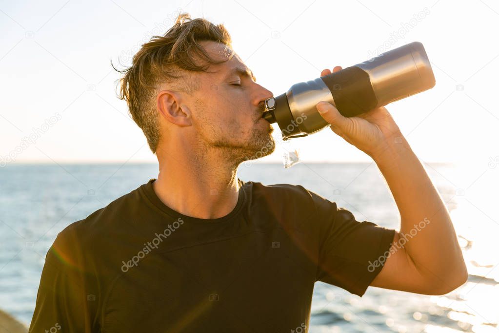 side view of handsome adult man drinking water from fitness bottle on seashore in front of sunrise