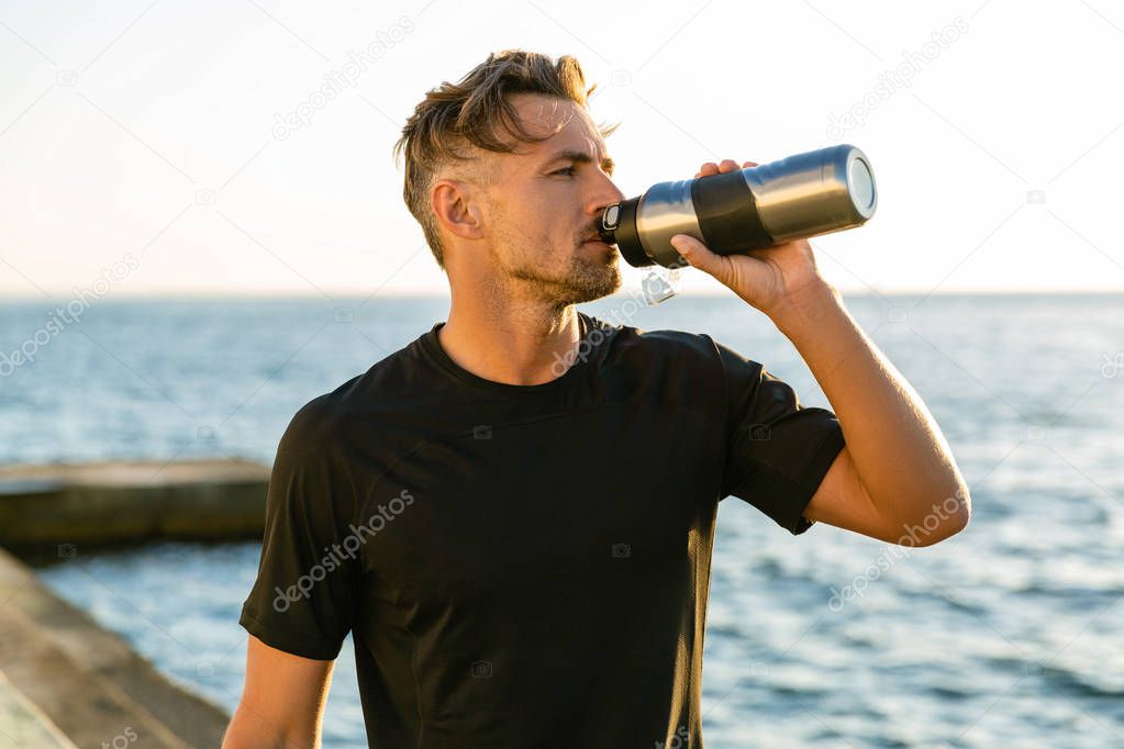 adult sportsman drinking water from fitness bottle on seashore in front of sunrise