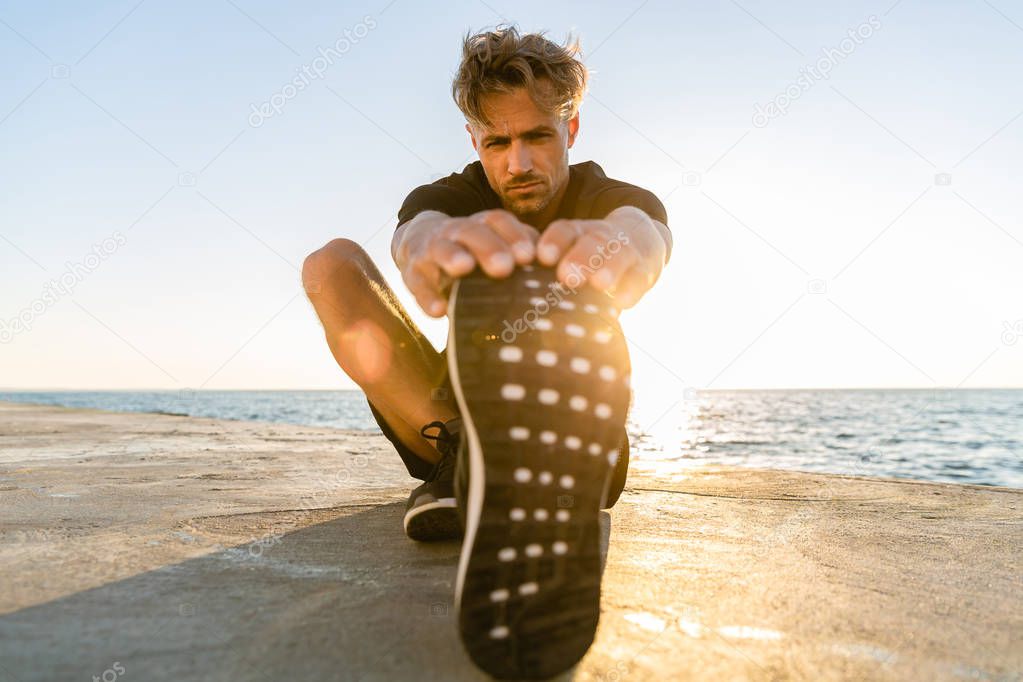 sporty adult man stretching leg on seashore in front of sunrise