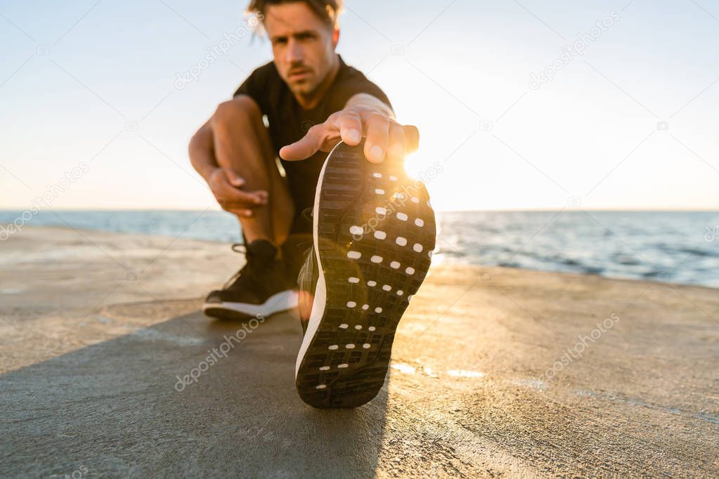 athletic adult man stretching leg on seashore in front of sunrise