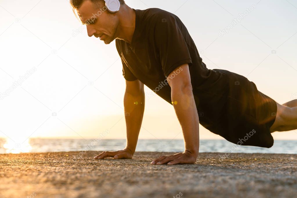 athletic adult man in headphones doing push ups on seashore