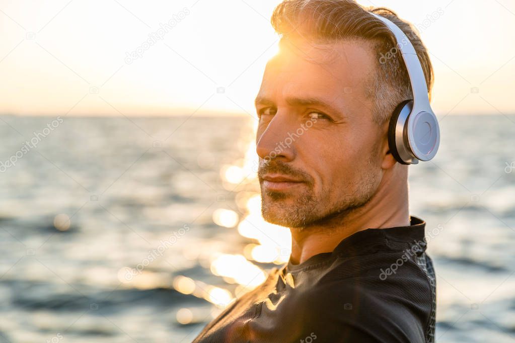 close-up portrait of smiling adult man in wireless headphones looking at camera on seashore in front of sunrise