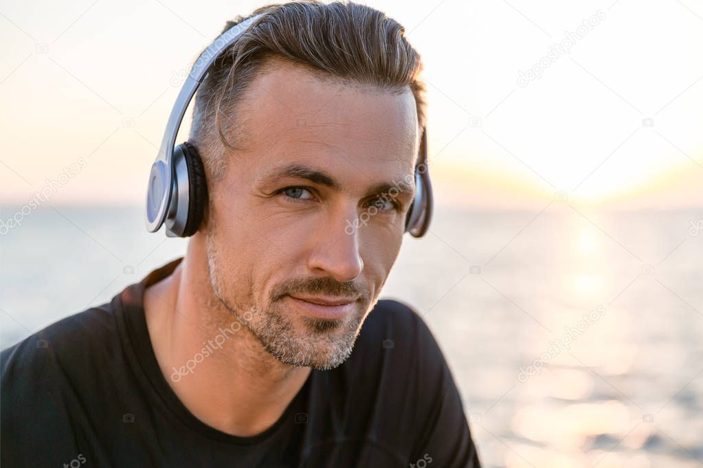 close-up portrait of attractive adult man in wireless headphones looking at camera on seashore
