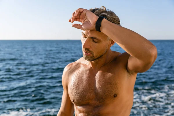 Müder, hemdloser erwachsener Mann wischt sich nach dem Training am Strand den Schweiß von der Stirn — Stockfoto