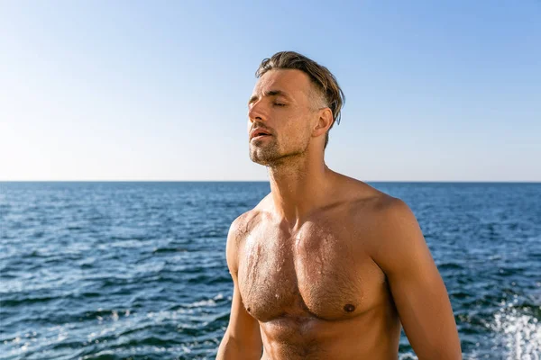 Sportif adulte mouillé torse nu avec les yeux fermés debout sur le bord de la mer — Photo de stock