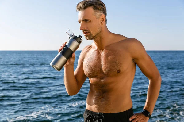 Shirtless handsome adult sportsman with fitness bottle looking away on seashore — Stock Photo