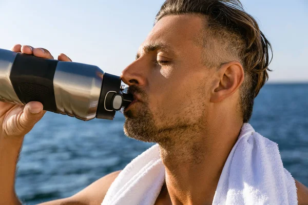 Close-up shot of shirtless handsome adult man with towel on shoulders drinking water after workout on seashore — Stock Photo