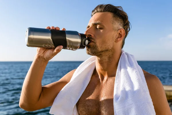 Hemdlos schöner erwachsener Sportler mit Handtuch auf den Schultern, der nach dem Training am Strand Wasser trinkt — Stockfoto