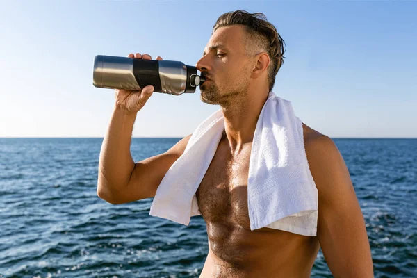 Sin camisa apuesto deportista adulto con toalla en los hombros beber agua después de entrenar en la orilla del mar - foto de stock