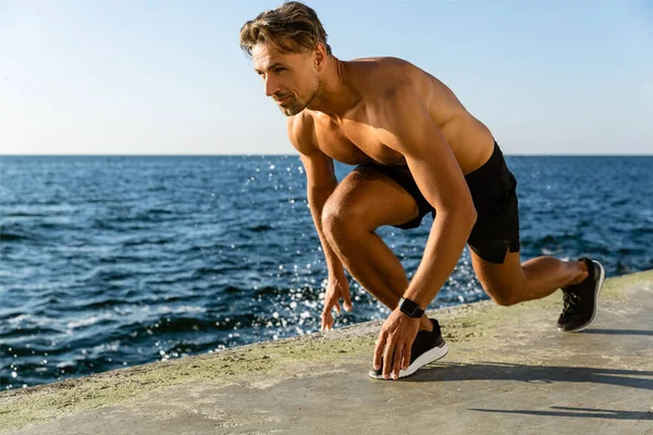 Apuesto adulto sin camisa corredor de sprint de pie en posición de inicio para correr en la orilla del mar - foto de stock