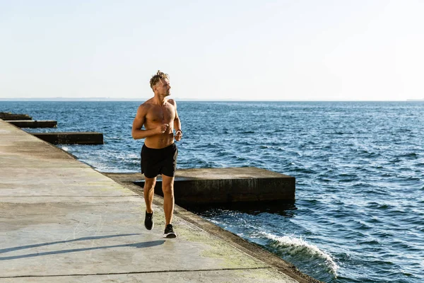 Deportista adulto sin camisa hombre trotando en la orilla del mar - foto de stock