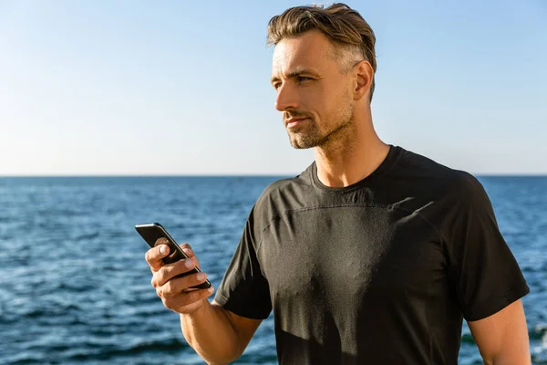 Handsome adult man with smartphone on seashore and looking away — Stock Photo