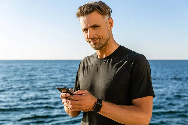 Homem adulto bonito com smartphone na praia e olhando para a câmera — Fotografia de Stock