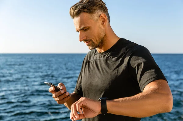Handsome adult man using smartphone and smart watch on seashore — Stock Photo