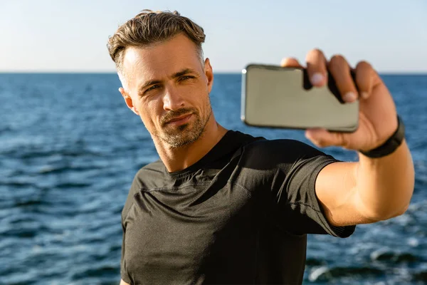 Guapo hombre adulto tomando selfie con teléfono inteligente en la orilla del mar - foto de stock