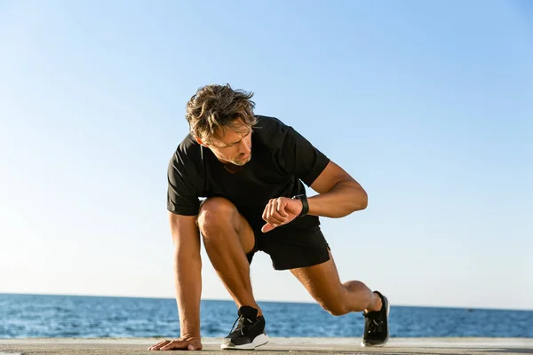 Apuesto corredor de sprint adulto mirando el rastreador de fitness mientras está de pie en la posición de inicio para correr en la orilla del mar - foto de stock