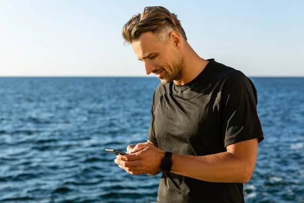 Homme adulte souriant utilisant un smartphone sur le bord de la mer — Photo de stock