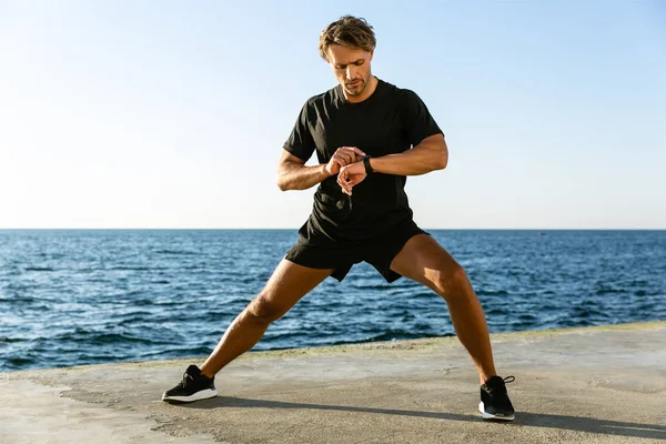 Handsome adult sportsman looking at fitness tracker while stretching before training on seashore — Stock Photo
