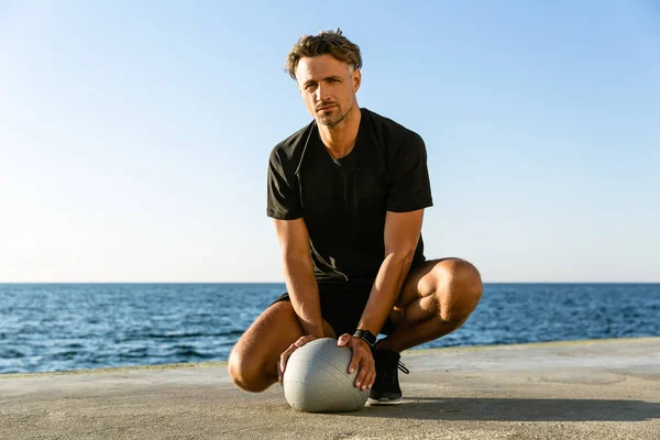 Bell'uomo adulto seduto con palla in forma sulla riva del mare e guardando la fotocamera — Foto stock