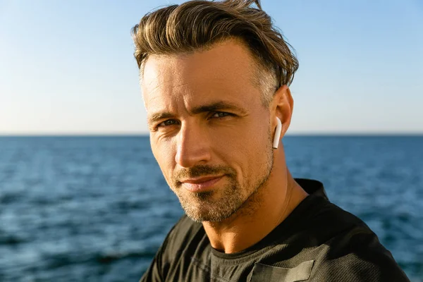 Retrato close-up de homem adulto com fones de ouvido sem fio na praia olhando para a câmera — Fotografia de Stock