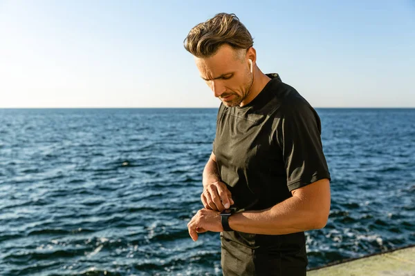 Schöner erwachsener Mann mit drahtlosen Kopfhörern, der die Trainingsergebnisse mit intelligenter Uhr am Meeresufer überprüft — Stockfoto