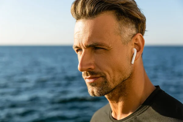 Portrait en gros plan d'un homme adulte attrayant avec écouteurs sans fil sur le bord de la mer — Photo de stock